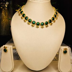 Gold-toned traditional necklace and earring set with round emerald-green stones and small red accent beads, displayed on an off-white velvet bust – South Indian Kemp-style temple jewellery by Santosree Collective
