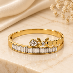 Gold-toned bangle bracelet with baguette crystal bar, pear-cut crystal and leaf motif detailing, laid on cream satin with dried flowers – anti-tarnish daily wear jewellery by Santosree Collective
