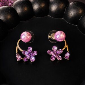 A pair of purple floral earrings with lavender pearl studs, gold-tone curved detailing, and purple crystal flower drops, placed on a black textured tray with purple dried flowers.