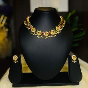 Traditional gold-tone necklace set displayed on a black jewellery stand, featuring red and green stone detailing, circular gold motifs, pearl drops, and matching stud earrings on both sides.