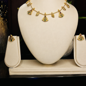 Antique gold-toned temple necklace and earring set with dangling bell-shaped charms, maroon and green stone accents, on a delicate chain, displayed on an off-white velvet bust – South Indian temple jewellery by Santosree Collective